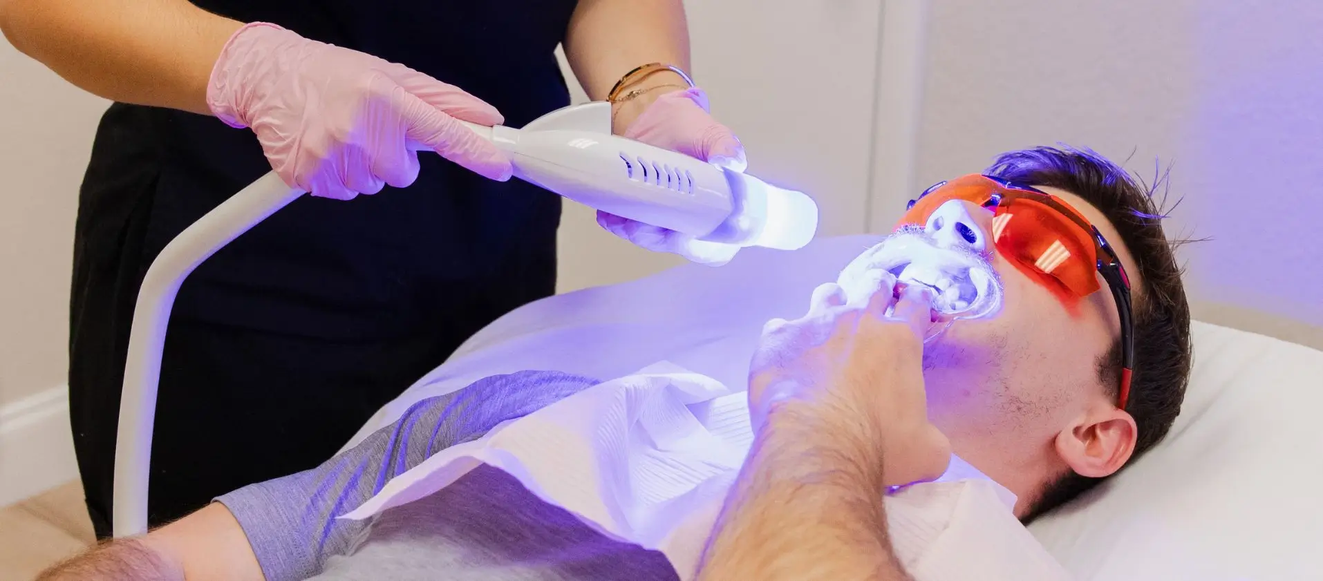 A person receives teeth whitening treatment while lying down, wearing protective glasses. Another person operates a UV light device.